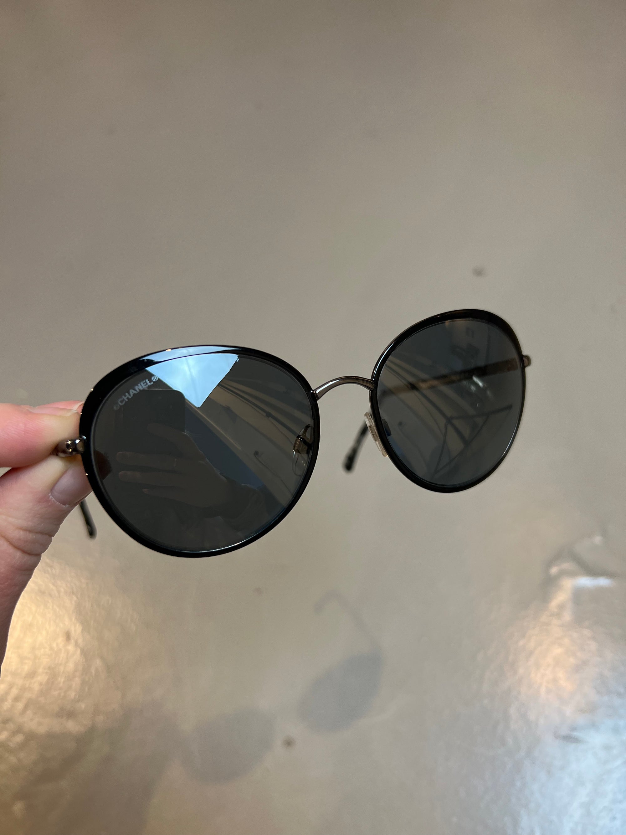 Detail-Produktbild einer runden Chanel Sonnenbrille in schwarz vor grauem Hintergrund.