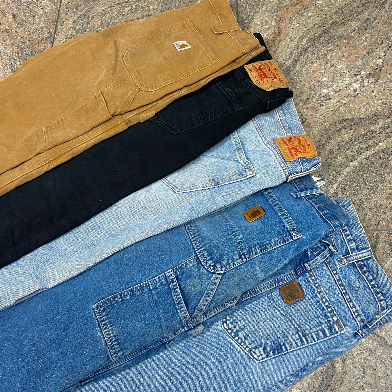 Zu sehen sind 5 verschiedene Jeans Hosen. Dabei vertreten sind Marken wie Carhartt. Die Hosen haben unterschiedliche Farben von Denim Blau, über Beige bis zu schwarz. 