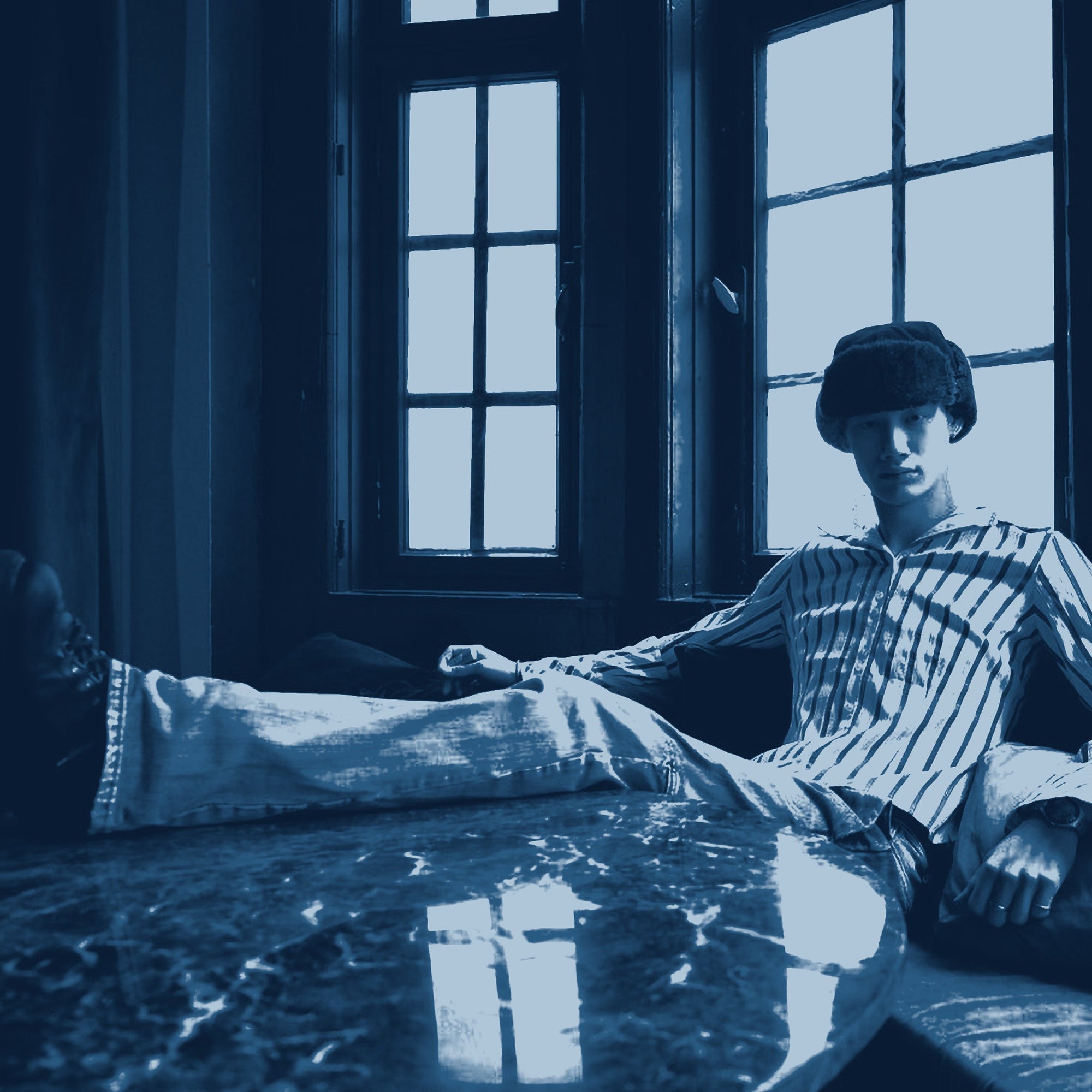 Zu sehen ist ein männliches Model. Die Person sitzt an einem Tisch, sie hat ein Bein auf dem Tisch und ist eingekleidet in unseren Vintage Klamotten.