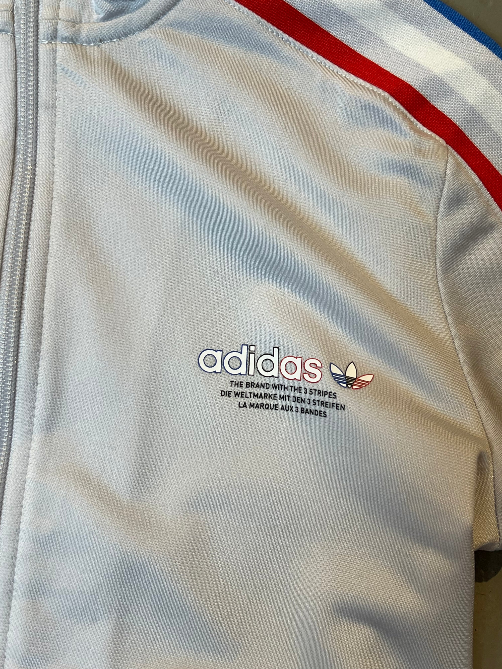 Produktbild von weißer Adidas Jacke Vorderseite mit roten und blauen Streifen und Logo auf der linken Brust. Nahaufnahme von Adidas Wort- und bildlogo.