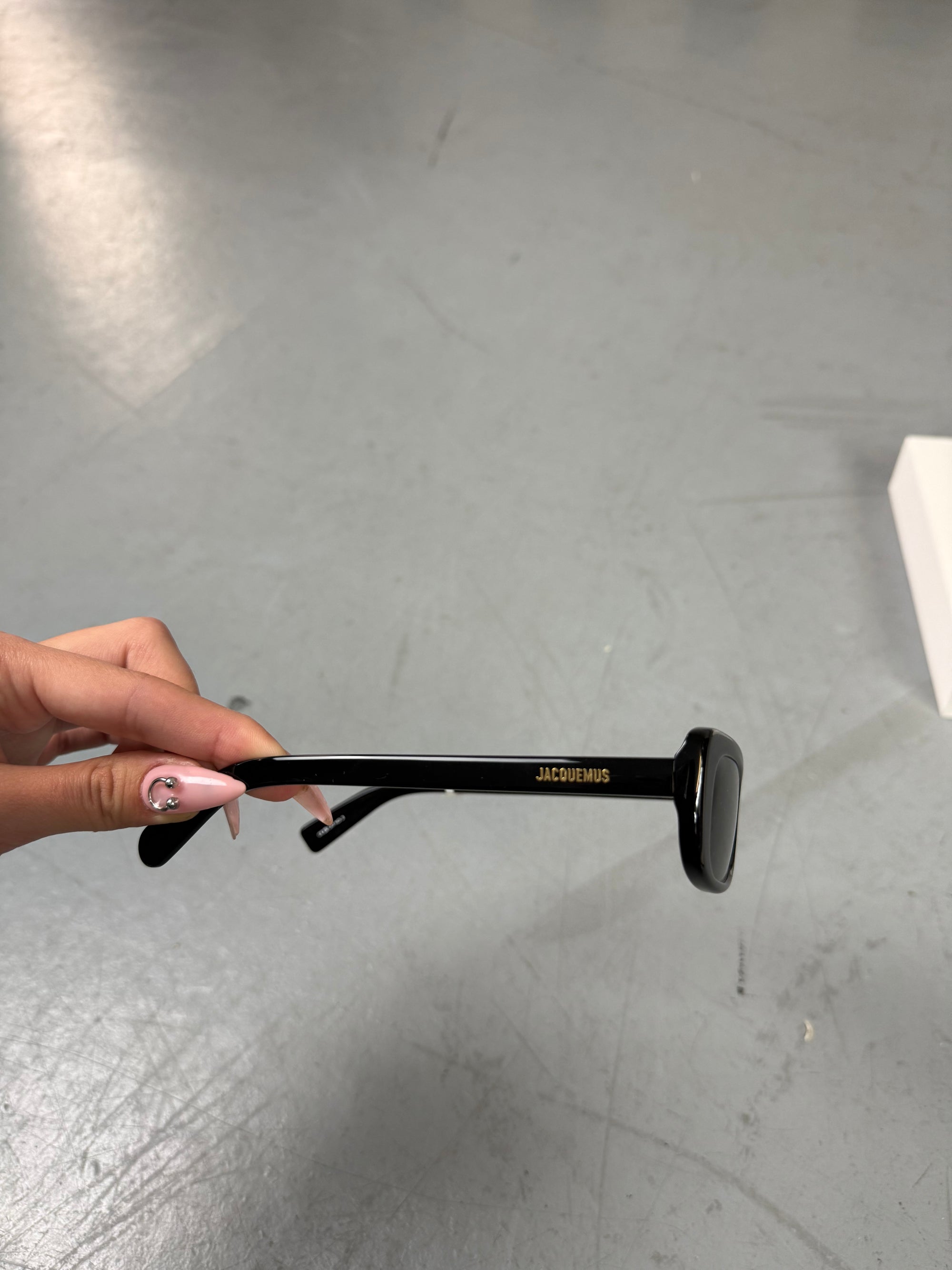 Eine schwarze schmale Sonnenbrille von Jacquemus mit goldenem Brand na,e auf den Bügeln 