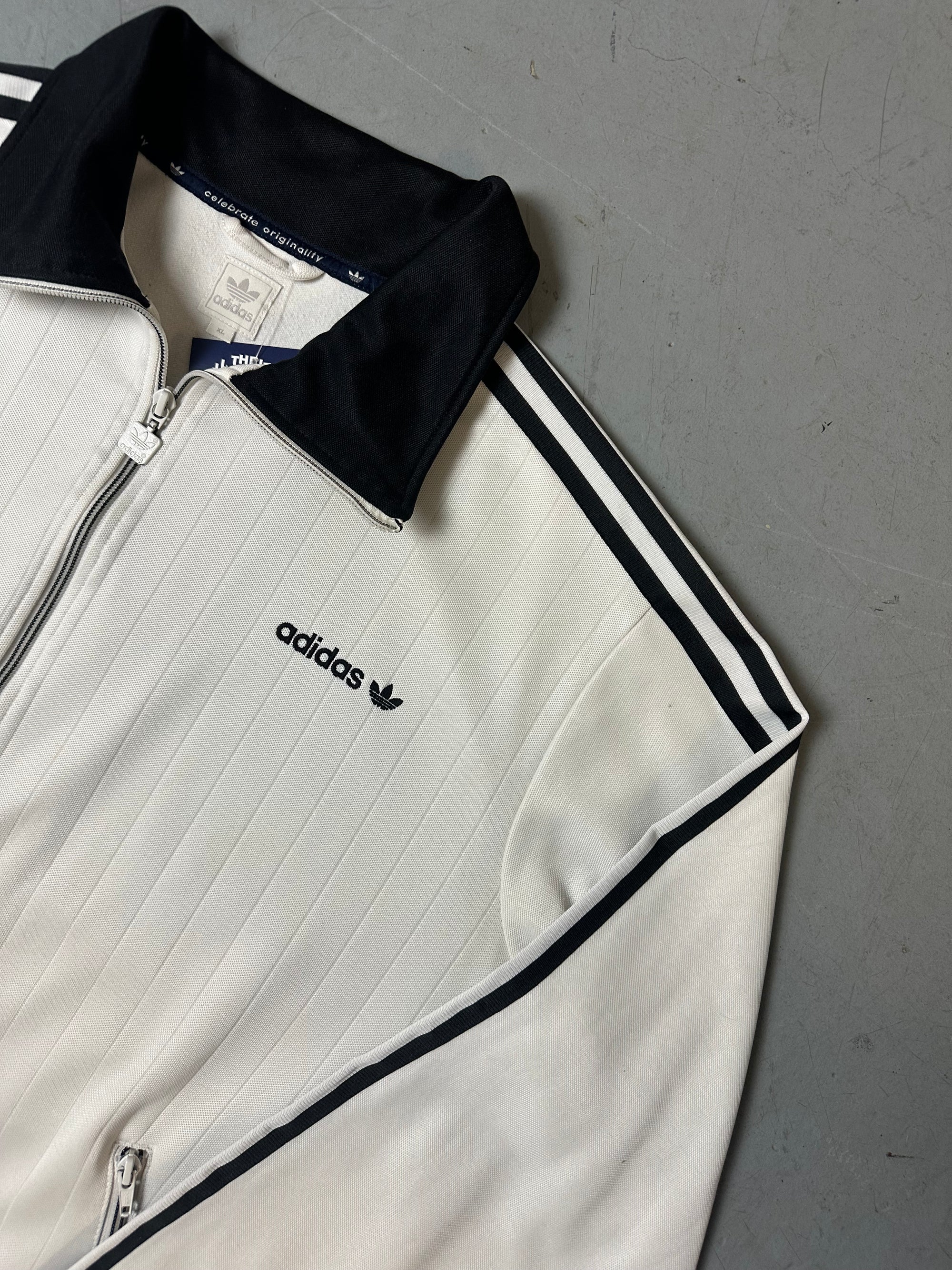 Eine weiße Trackjacke von Adidas mit schwarzen Streifen, schwarzer Kragen und dem Logo in schwarz auf der Brust 