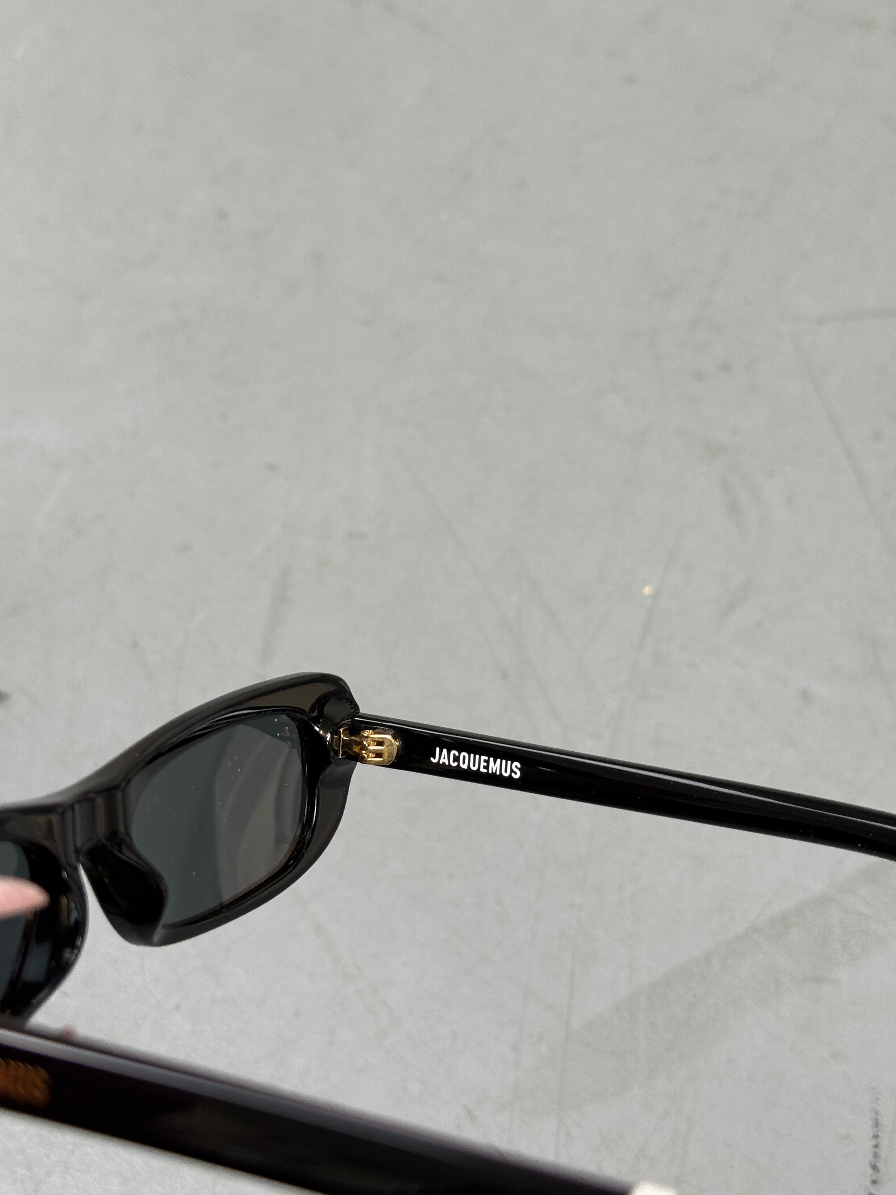 Eine schwarze schmale Sonnenbrille von Jacquemus mit goldenem Brand na,e auf den Bügeln 
