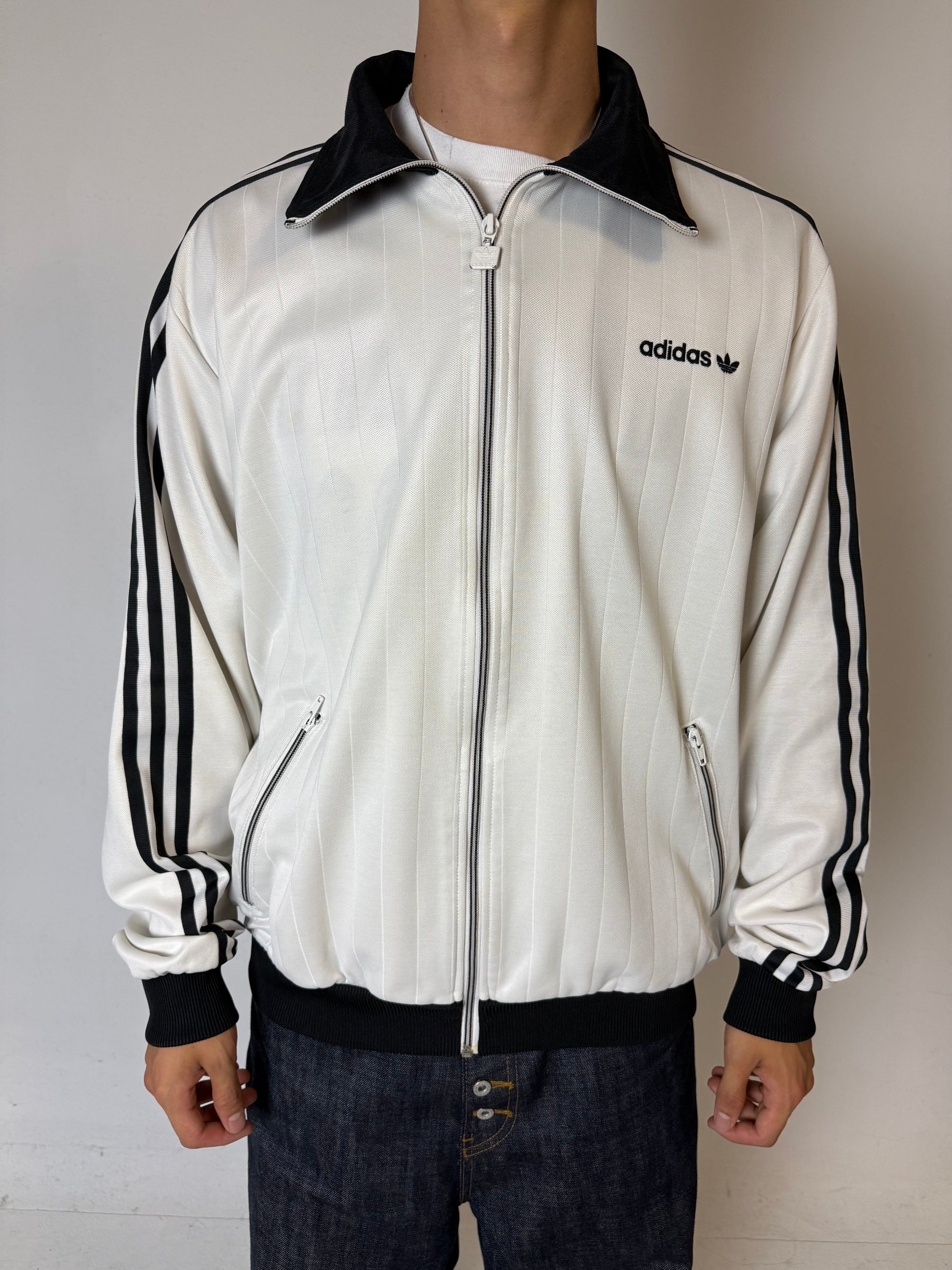 Eine weiße Trackjacke von Adidas mit schwarzen Streifen, schwarzer Kragen und dem Logo in schwarz auf der Brust 