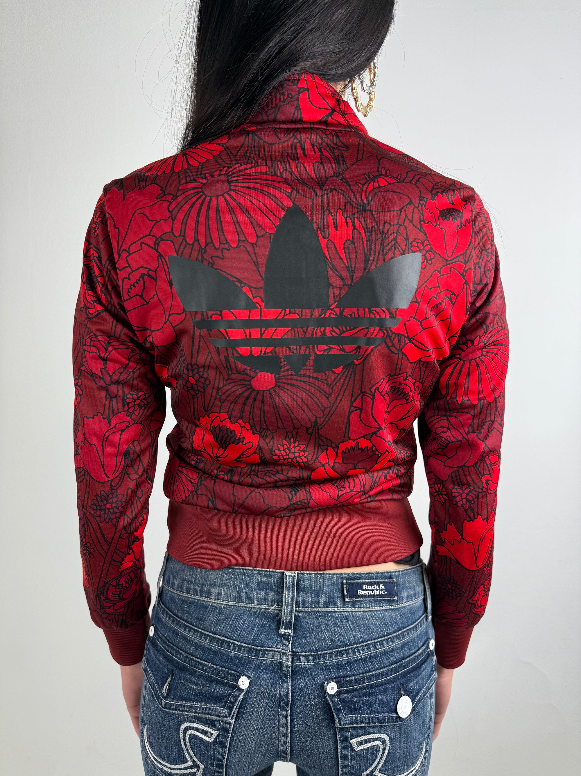 Frau trägt eine bordeauxrote Adidas-Trainingsjacke mit auffälligem roten Blumenmuster und schwarzen Streifen an den Ärmeln und einem Adidas Logo auf dem Rücken, kombiniert mit einer dunkelblauen Jeans. Die Jacke hat einen Reißverschluss, Rippbündchen und einen sportlich-femininen Look.