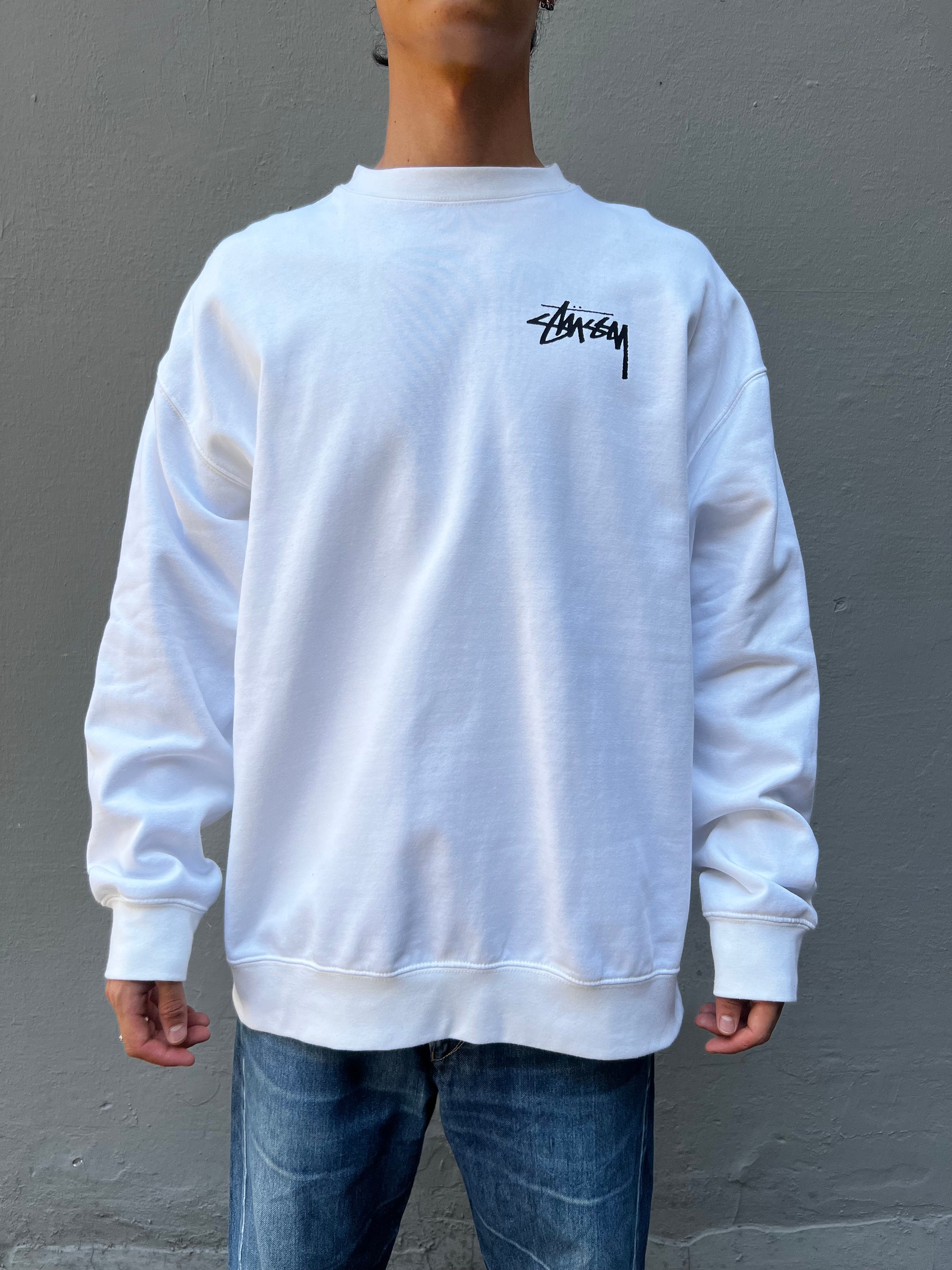 Tragebild von weißem Stüssy Longsleeve von vorne vor grauer Wand