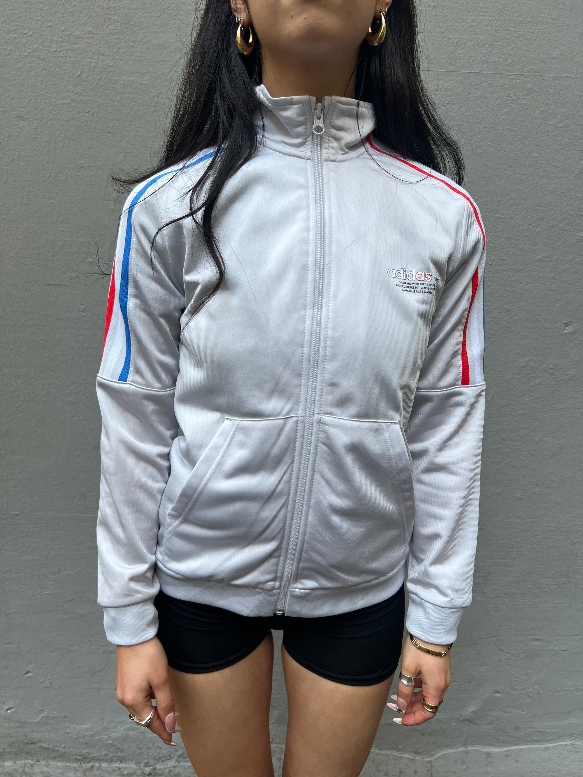 Tragebild von weißer Adidas Trainingsjacke mit roten und blauen Streifen und Adidas Logo auf der linken Brust