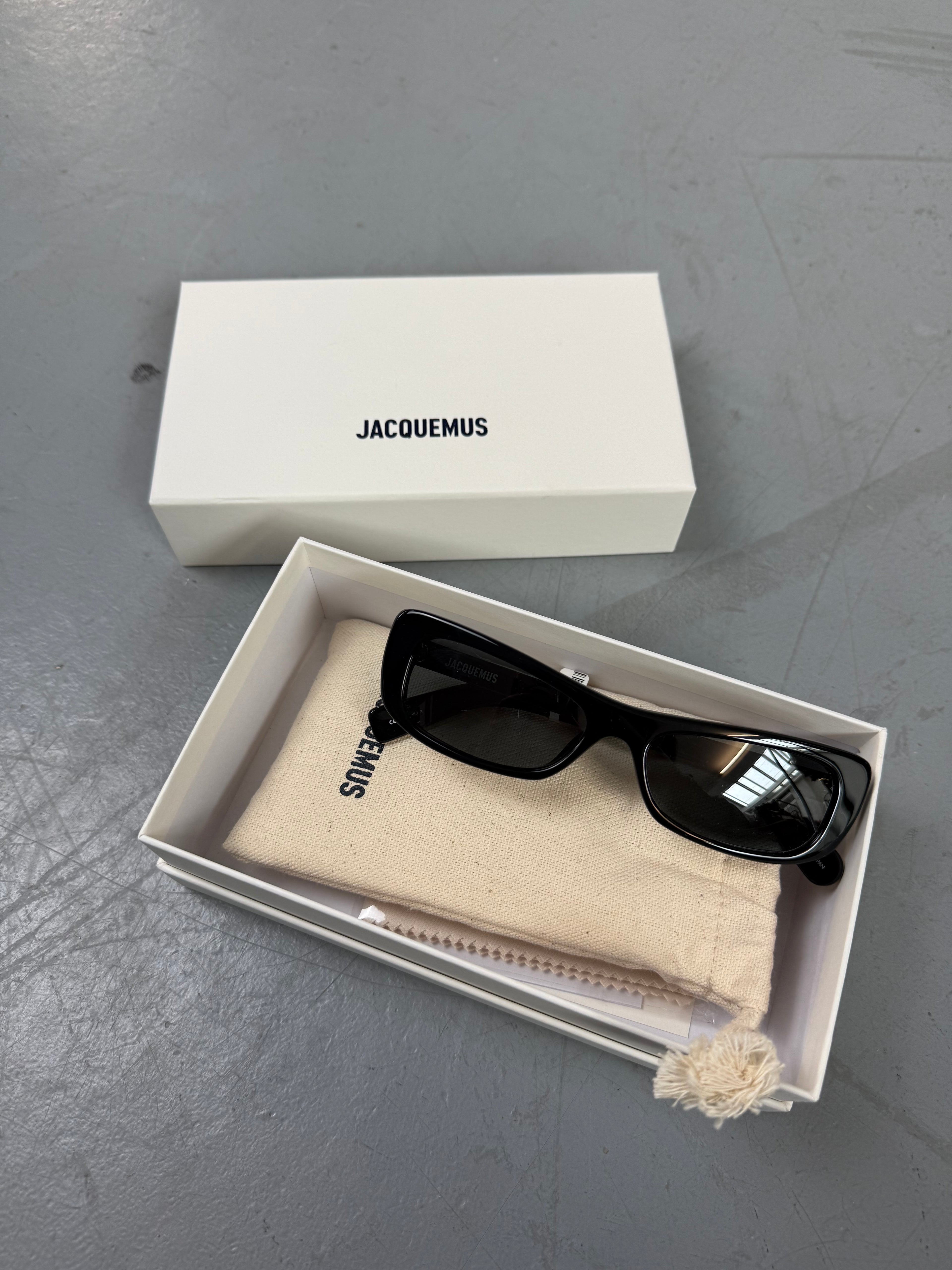 Eine schwarze schmale Sonnenbrille von Jacquemus mit goldenem Brand na,e auf den Bügeln 