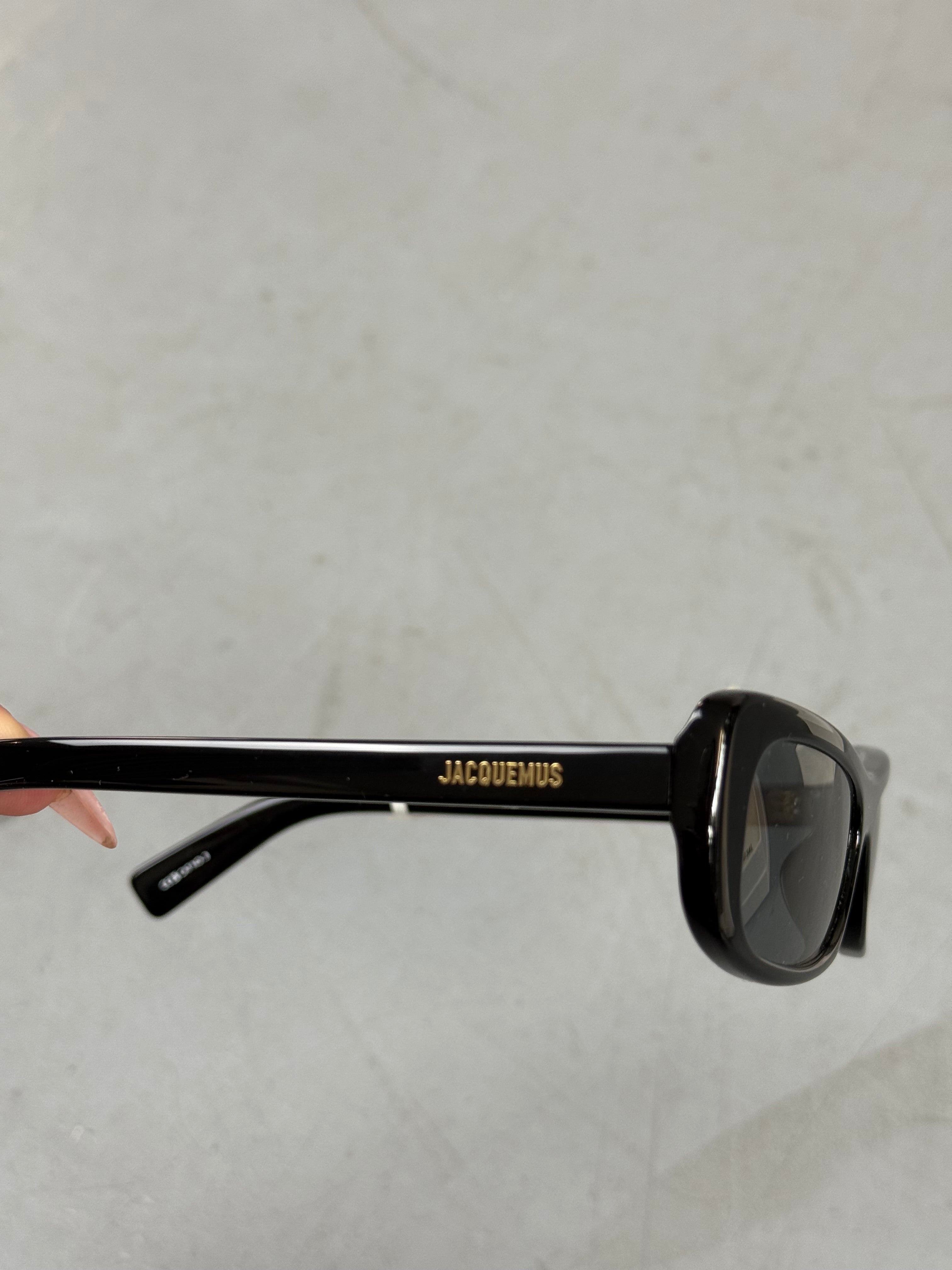 Eine schwarze schmale Sonnenbrille von Jacquemus mit goldenem Brand na,e auf den Bügeln 