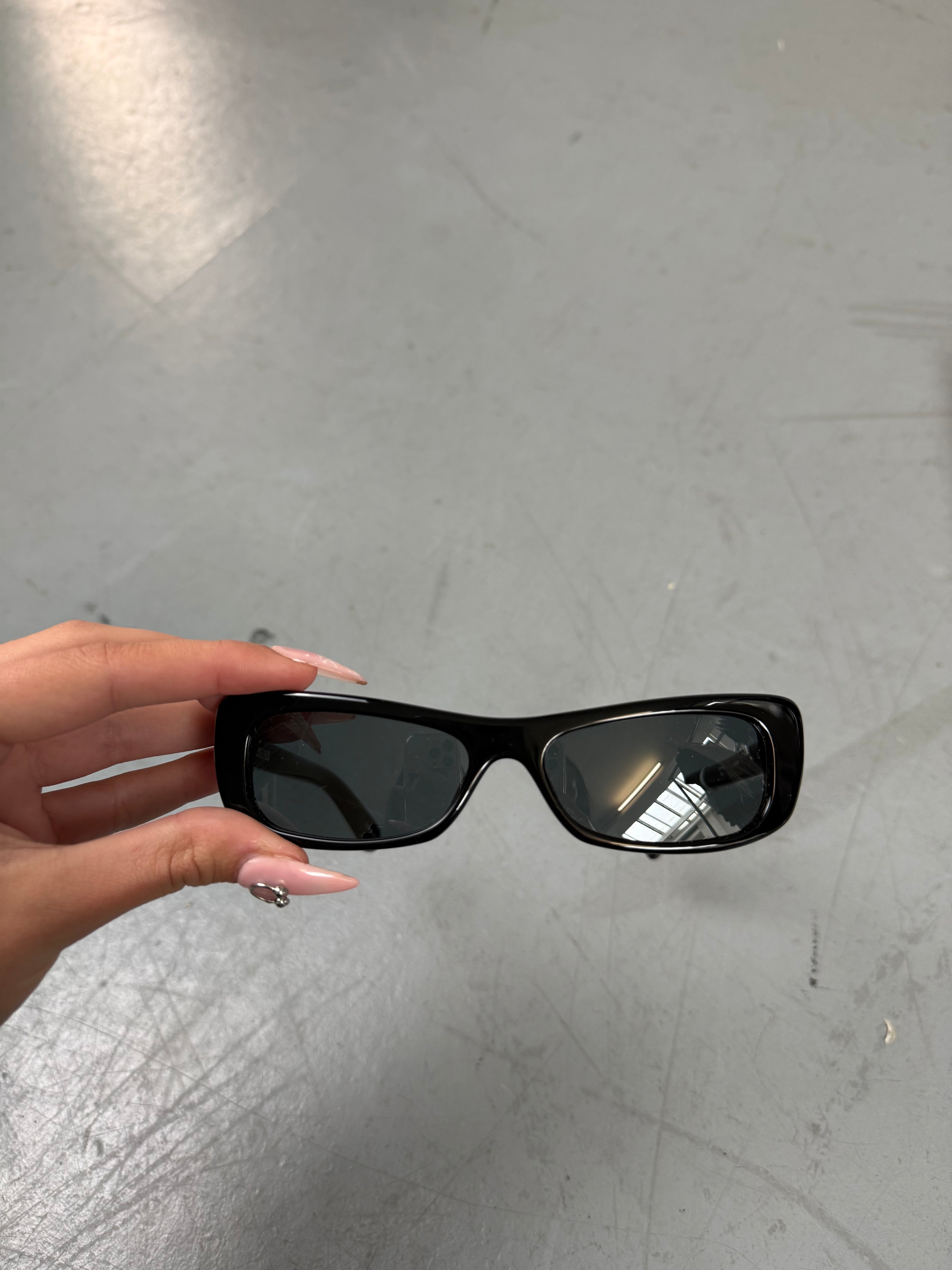 Eine schwarze schmale Sonnenbrille von Jacquemus mit goldenem Brand na,e auf den Bügeln 