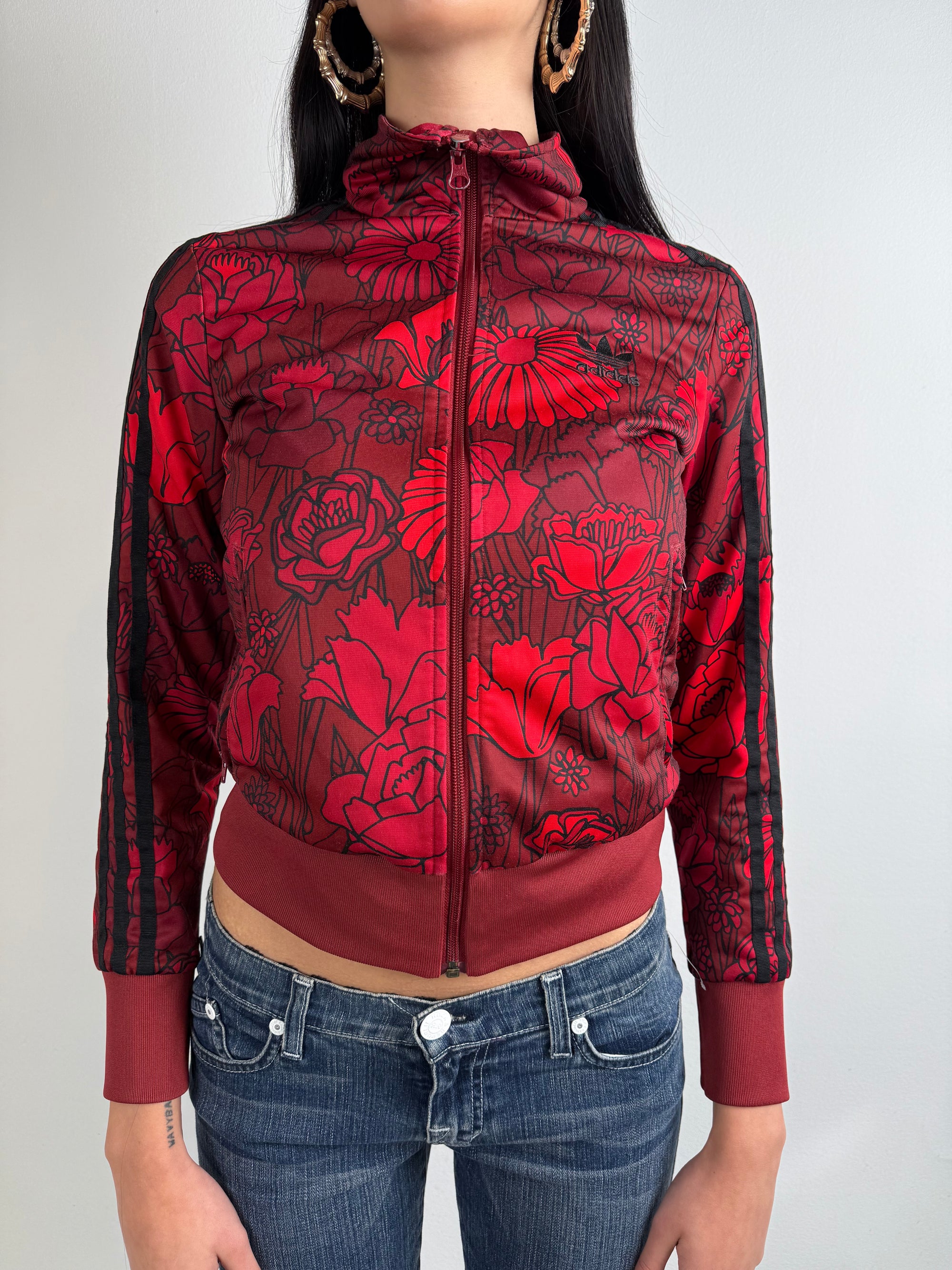 Frau trägt eine bordeauxrote Adidas-Trainingsjacke mit auffälligem roten Blumenmuster und schwarzen Streifen an den Ärmeln, kombiniert mit einer dunkelblauen Jeans. Die Jacke hat einen Reißverschluss, Rippbündchen und einen sportlich-femininen Look.
