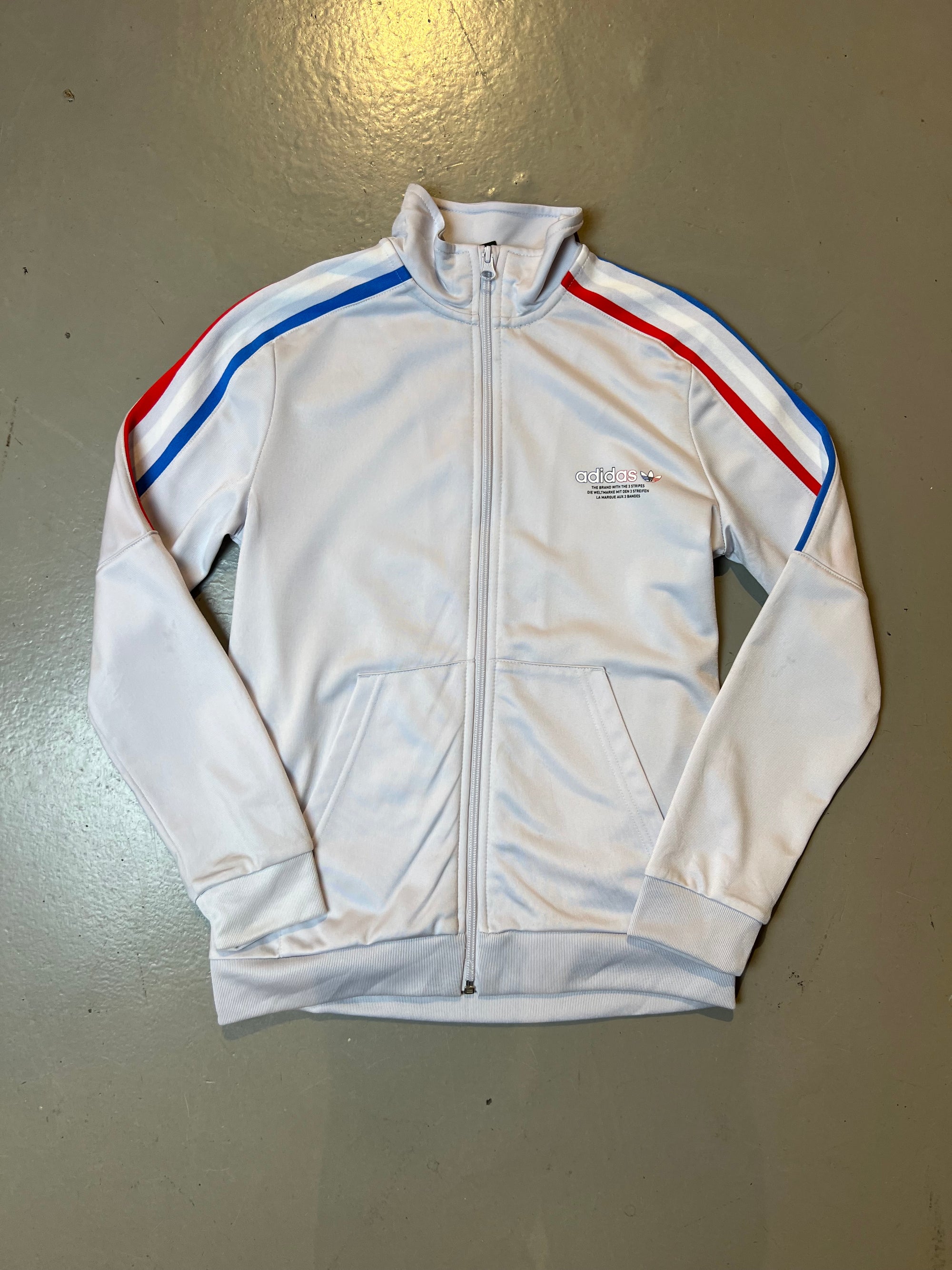 Produktbild von weißer Adidas Jacke Vorderseite mit roten und blauen Streifen und Logo auf der linken Brust.
