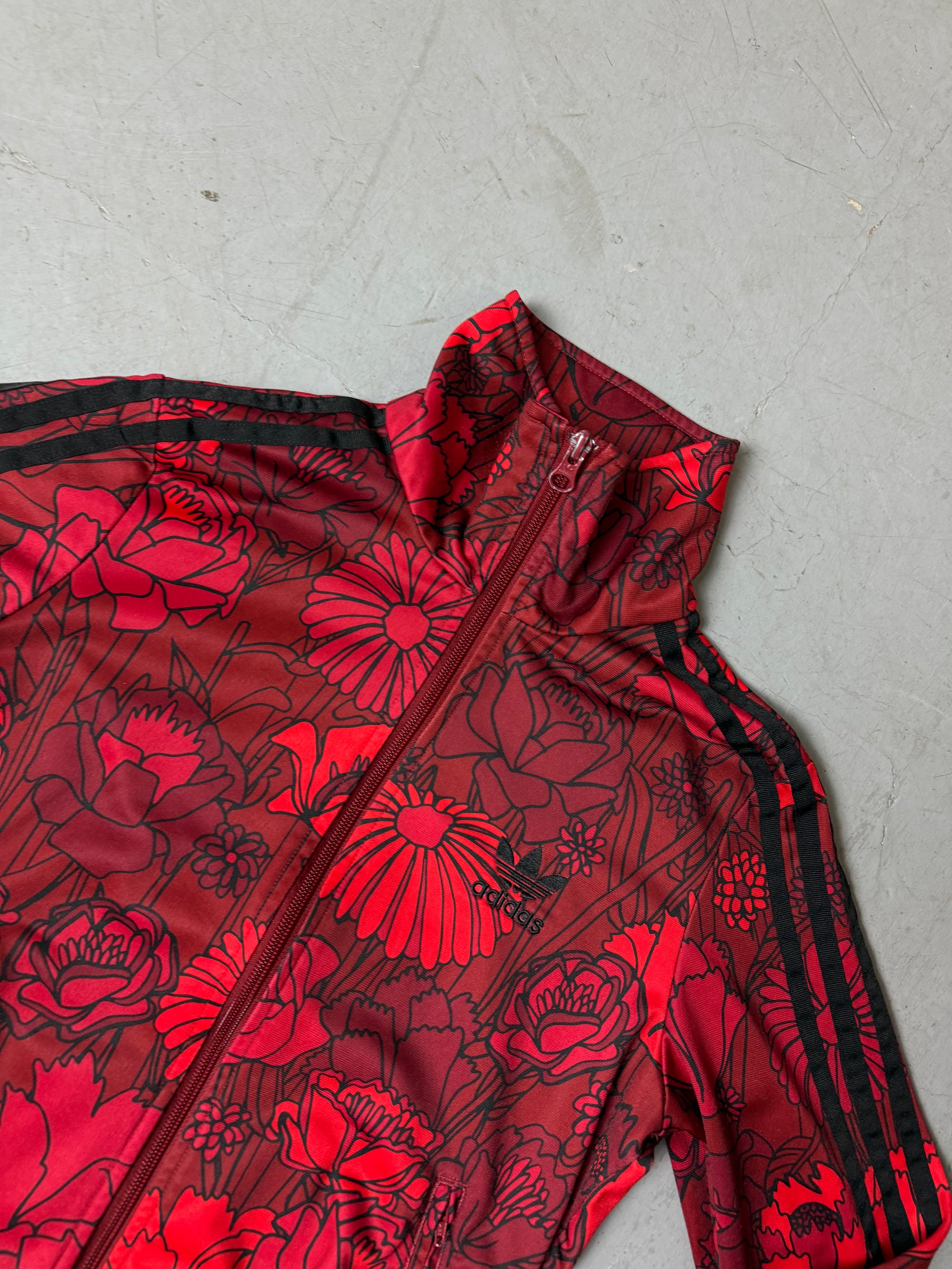 Frau trägt eine bordeauxrote Adidas-Trainingsjacke mit auffälligem roten Blumenmuster und schwarzen Streifen an den Ärmeln, kombiniert mit einer dunkelblauen Jeans. Die Jacke hat einen Reißverschluss, Rippbündchen und einen sportlich-femininen Look.