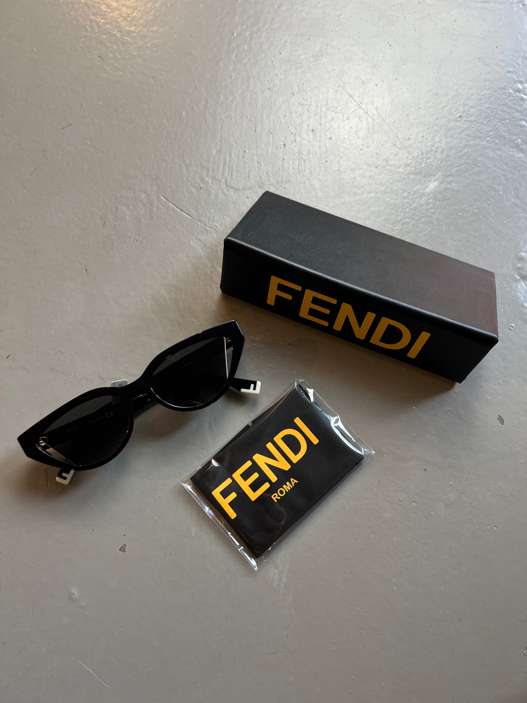 Produktbild von schwarzer Fendi Cat-Eye Sonnenbrille vor grauem Hintergrund. Die schwarz-gelbe Hülle ist beigelegt.