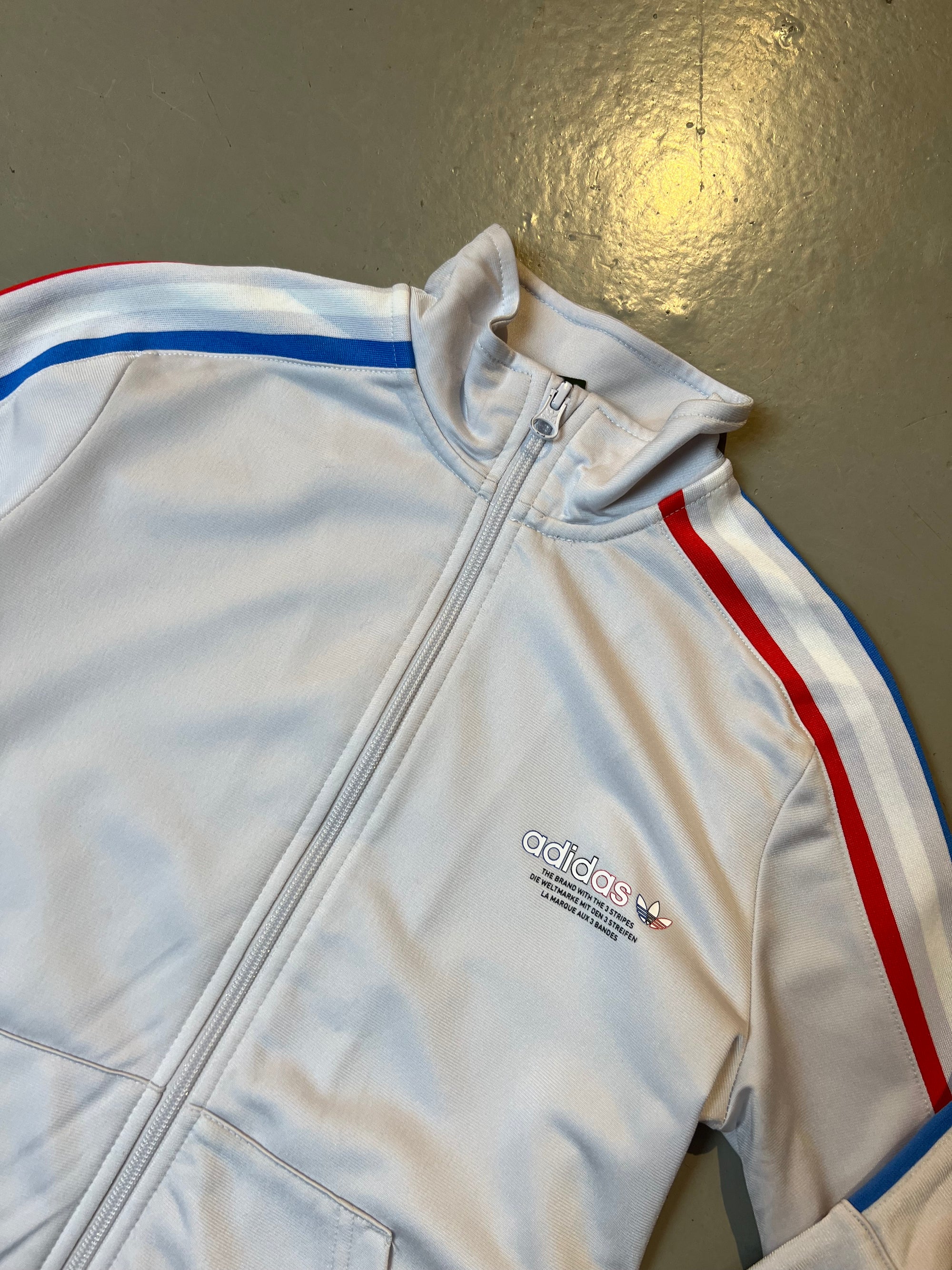 Produktbild von weißer Adidas Jacke Vorderseite mit roten und blauen Streifen und Logo auf der linken Brust.