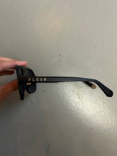 Laden Sie das Bild in den Galerie-Viewer, Zu sehen ist ein Derailbild von der Aufschrift auf dem Brillengestell einer Philipp Plein Sunglasses von der Seite vor einem grauen Hintergrund.