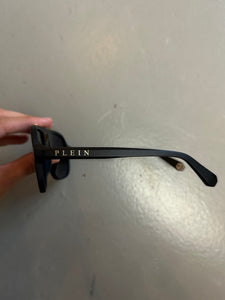 Zu sehen ist ein Derailbild von der Aufschrift auf dem Brillengestell einer Philipp Plein Sunglasses von der Seite vor einem grauen Hintergrund.