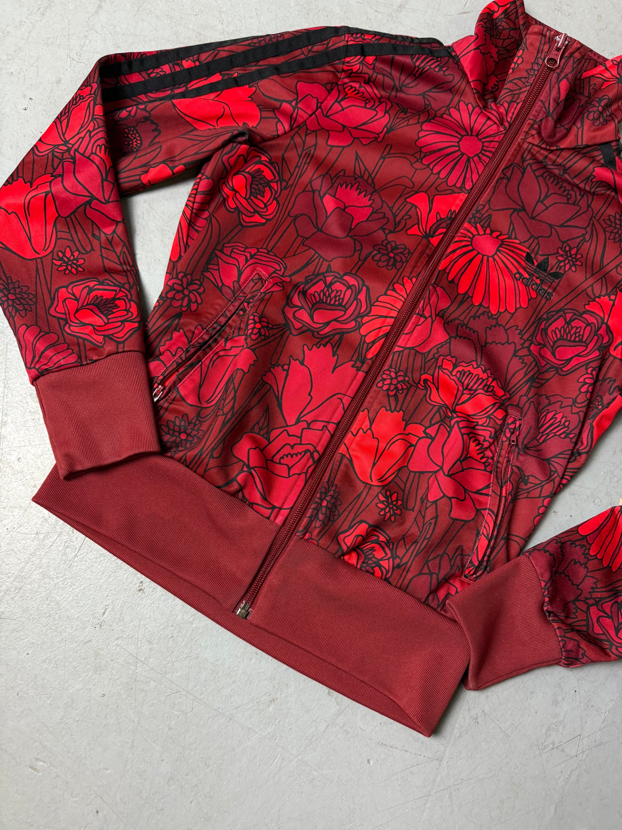 Frau trägt eine bordeauxrote Adidas-Trainingsjacke mit auffälligem roten Blumenmuster und schwarzen Streifen an den Ärmeln, kombiniert mit einer dunkelblauen Jeans. Die Jacke hat einen Reißverschluss, Rippbündchen und einen sportlich-femininen Look.