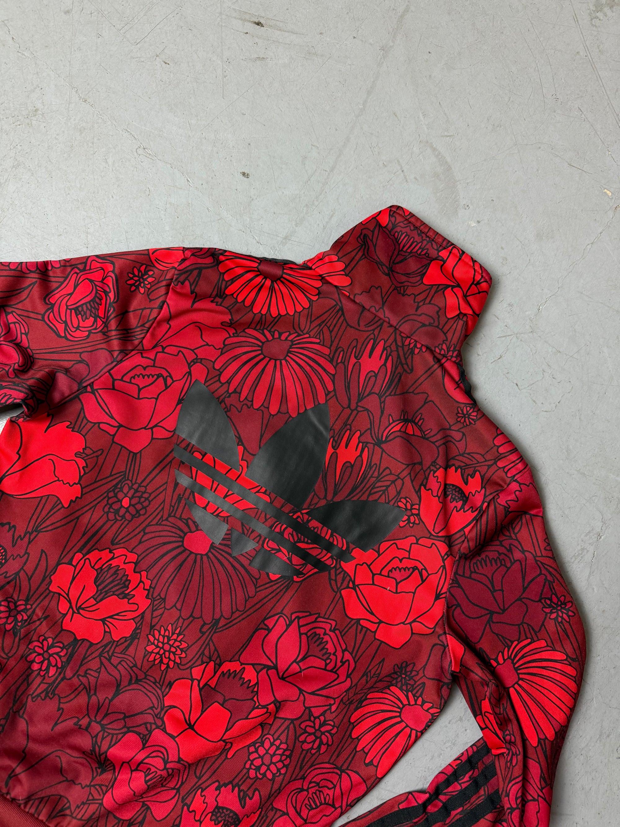 Frau trägt eine bordeauxrote Adidas-Trainingsjacke mit auffälligem roten Blumenmuster und schwarzen Streifen an den Ärmeln, kombiniert mit einer dunkelblauen Jeans. Die Jacke hat einen Reißverschluss, Rippbündchen und einen sportlich-femininen Look.