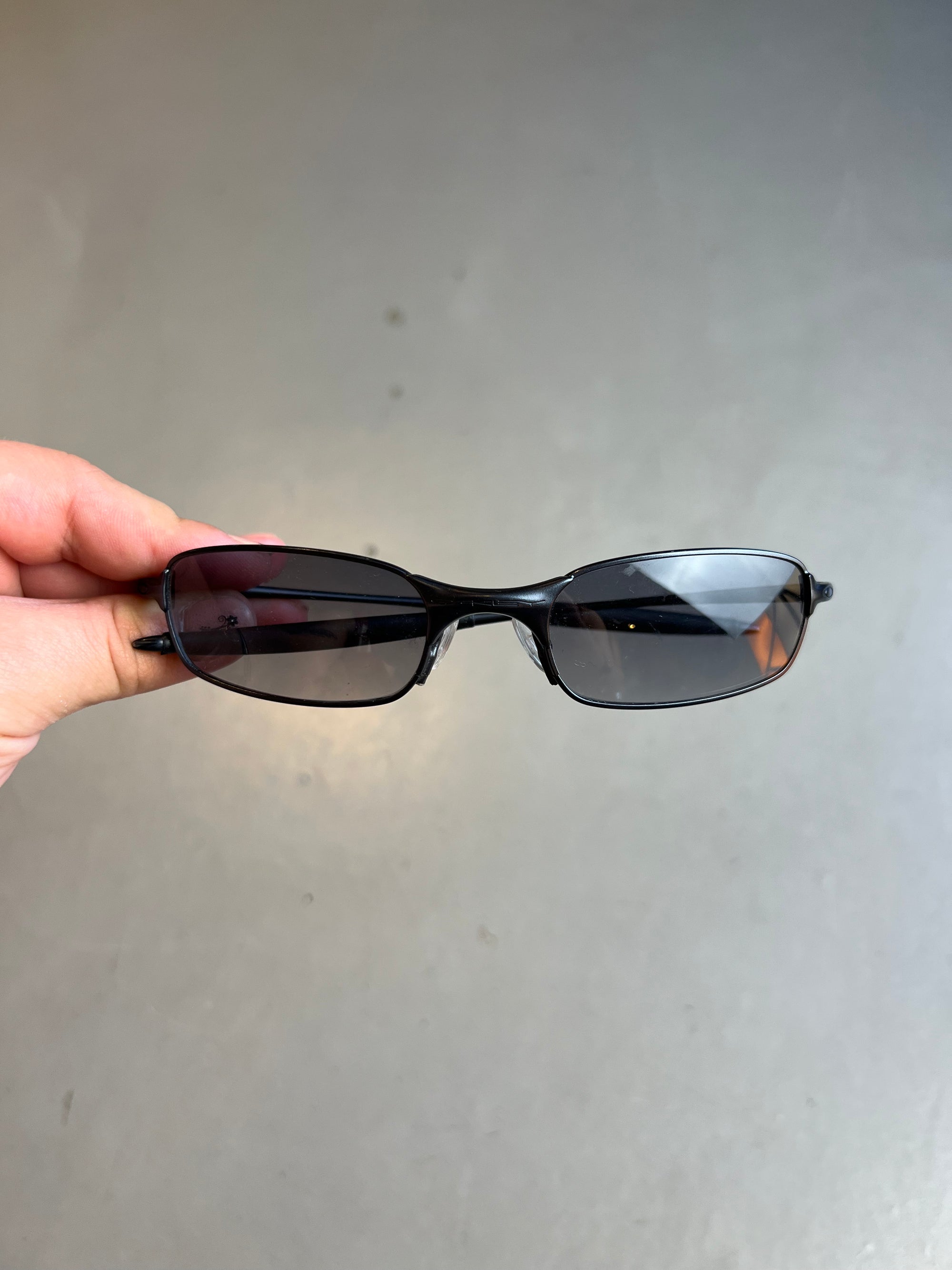 Auf dem Bild ist ein Produktbild der Oakley Raver Sunglasses in Schwarz von vorne vor einem grauem Hintergrund zu sehen.