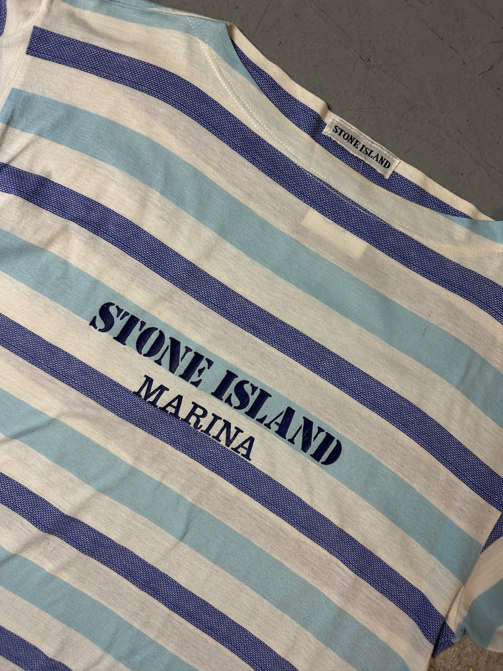 Detailliertes Produktbild von blau weis gestreiftem Stone Island Tshirt auf grauem Boden 