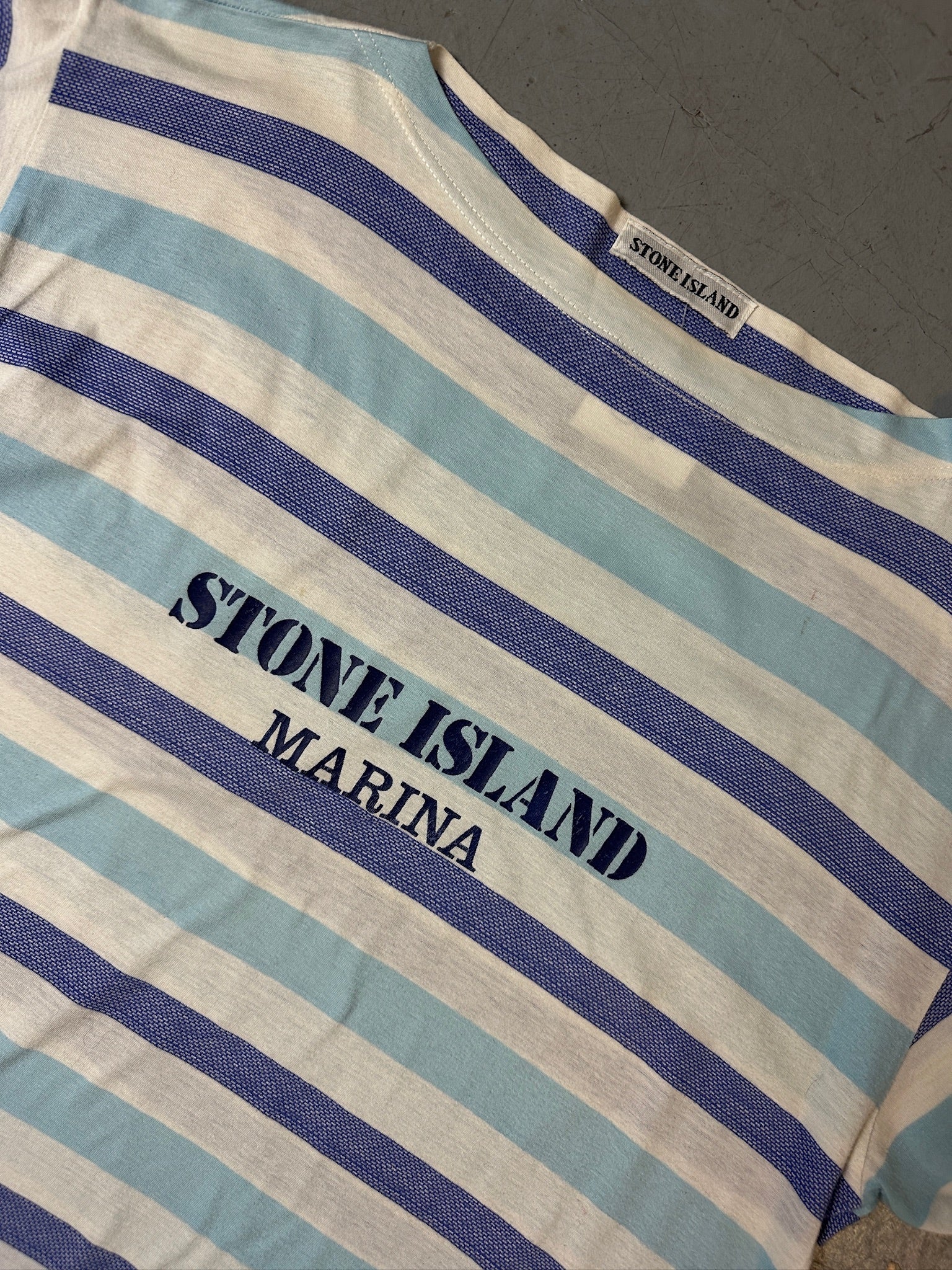 Detailliertes Produktbild von blau weis gestreiftem Stone Island Tshirt auf grauem Boden 