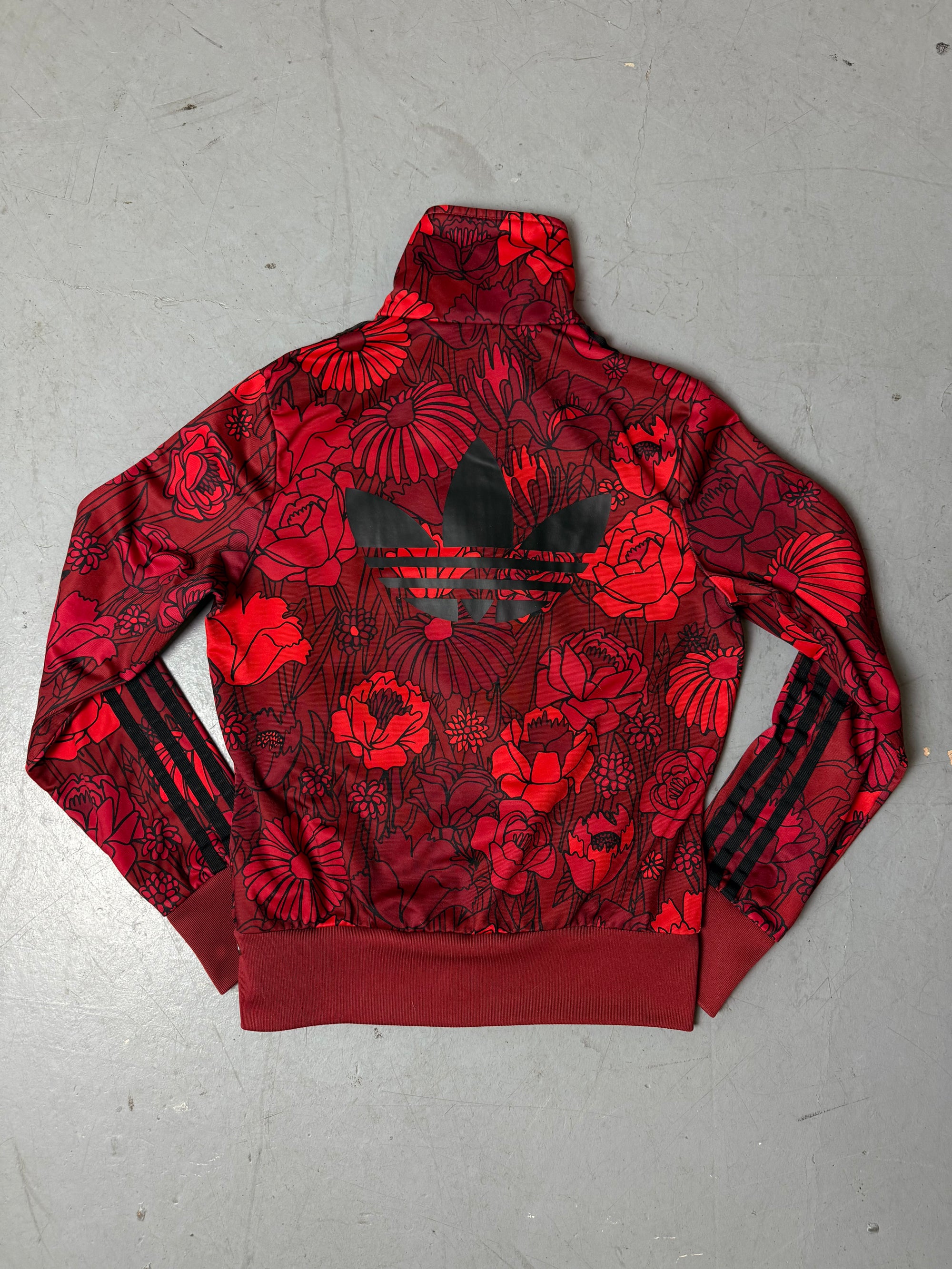 Frau trägt eine bordeauxrote Adidas-Trainingsjacke mit auffälligem roten Blumenmuster und schwarzen Streifen an den Ärmeln, kombiniert mit einer dunkelblauen Jeans. Die Jacke hat einen Reißverschluss, Rippbündchen und einen sportlich-femininen Look.