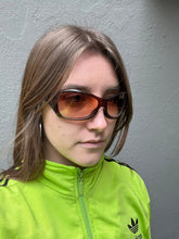 Laden Sie das Bild in den Galerie-Viewer, Tragebild von einer Braunen Oakley Sonnenbrille vor einer Grauen Wand