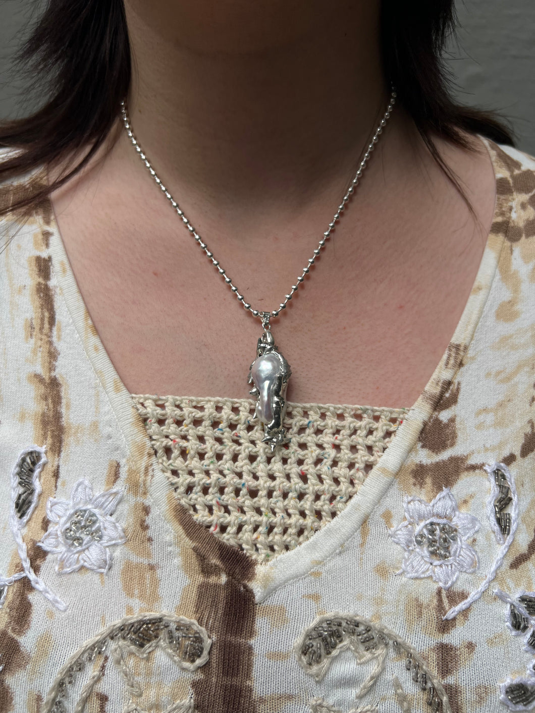 Tragebild von einer silbernen Perlenhalskette mit abstraktem Anhänger von Xullery vor einem grauen Hintergrund.