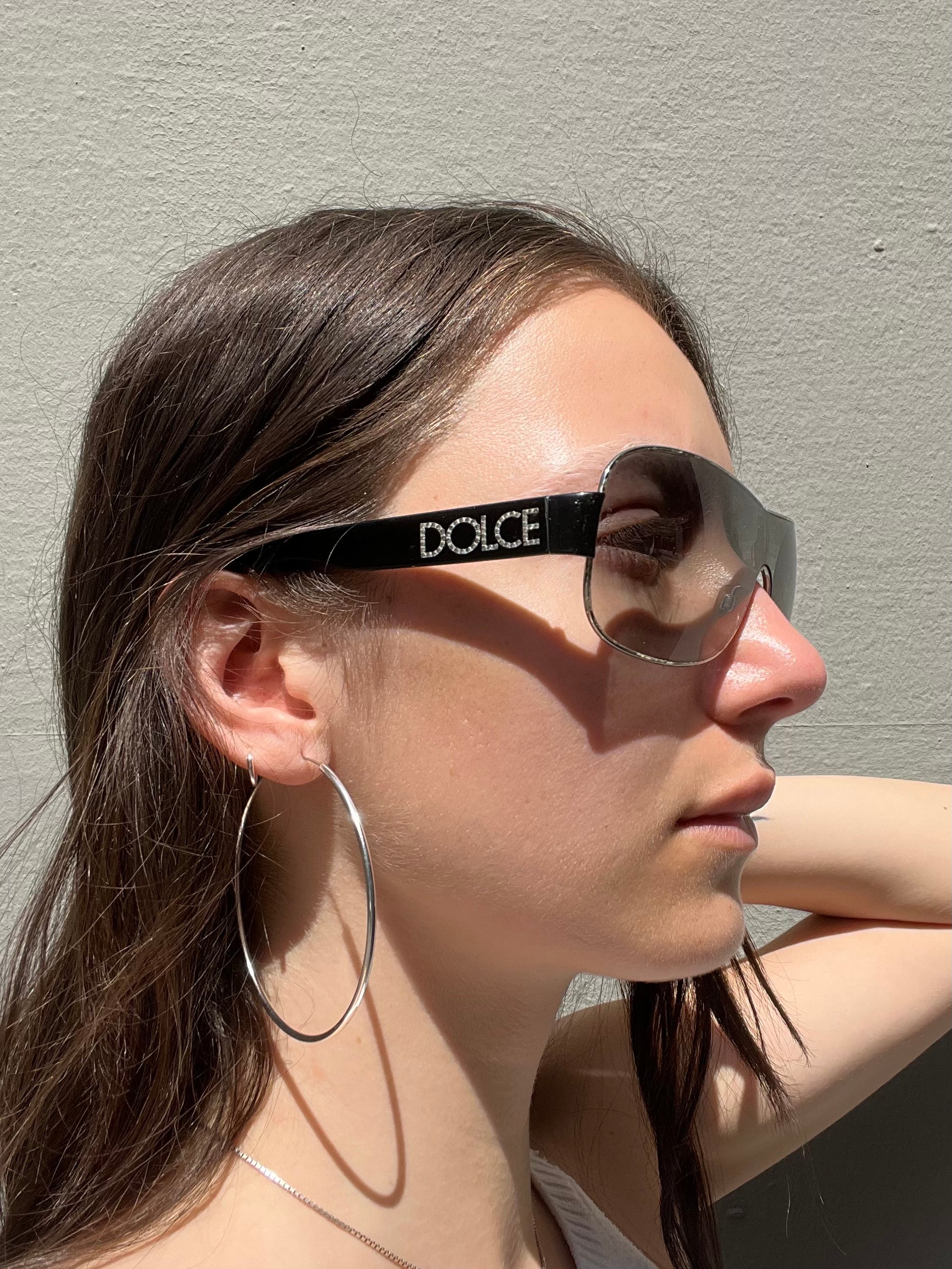Weibliches Model trägt Dolce&Gabbana Sonnenbrillle mit schwarzem Bügel und 2000er Stil vor grauer Wand.