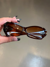 Laden Sie das Bild in den Galerie-Viewer, Produktbild von einer Braunen Oakley Sonnenbrille vor einer Grauen Wand