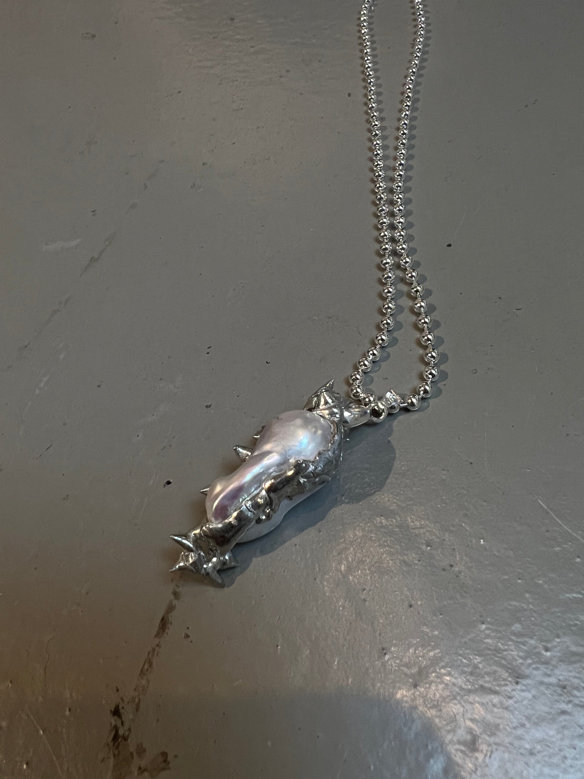 Produktbild von einer silbernen Perlenhalskette mit abstraktem Anhänger von Xullery vor einem grauen Hintergrund.