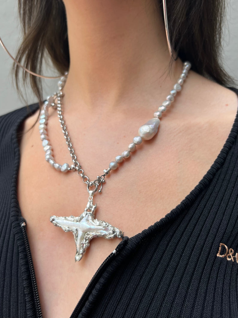 Tragebild von Xullery Cross Grey Pearl Necklace vor grauer Wand.