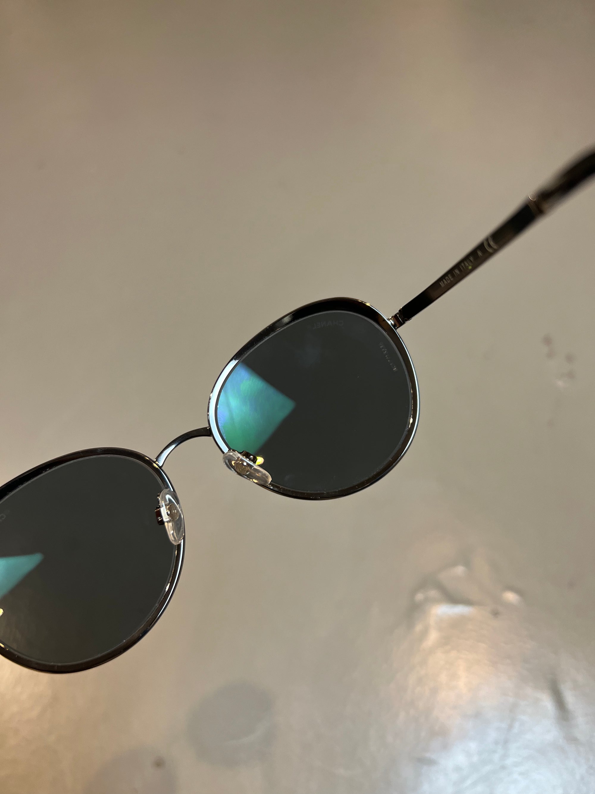 Detail-Produktbild einer runden Chanel Sonnenbrille in schwarz vor grauem Hintergrund.
